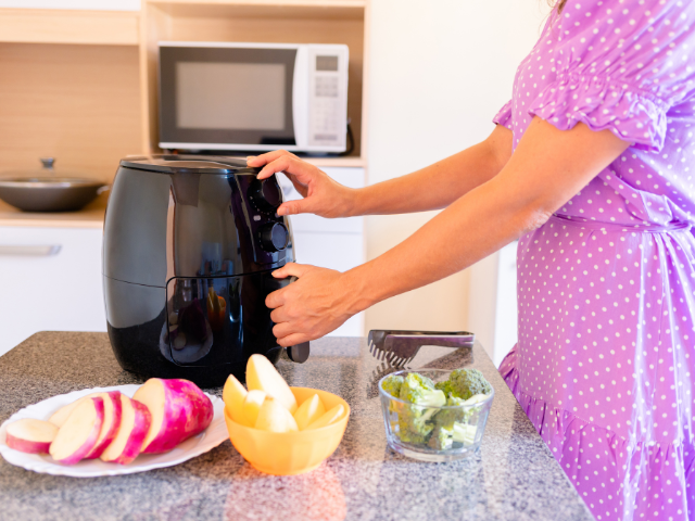 A woman in a purple polka-dot dress adjusts the settings of a black air fryer on a kitchen countertop, surrounded by fresh vegetables.