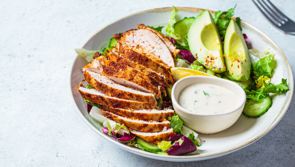 A fresh salad topped with seasoned chicken breast, avocado, and a side of creamy dressing for a wholesome meal.