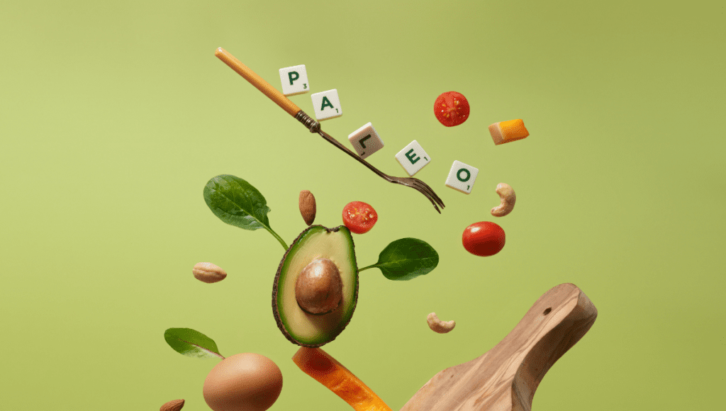 Paleo diet ingredients such as avocado, spinach, tomatoes, and a fork creatively floating against a green background.