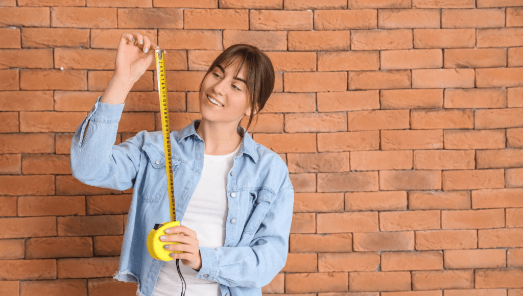 A young woman holding a yellow measuring tape and smiling in front of a brick wall.