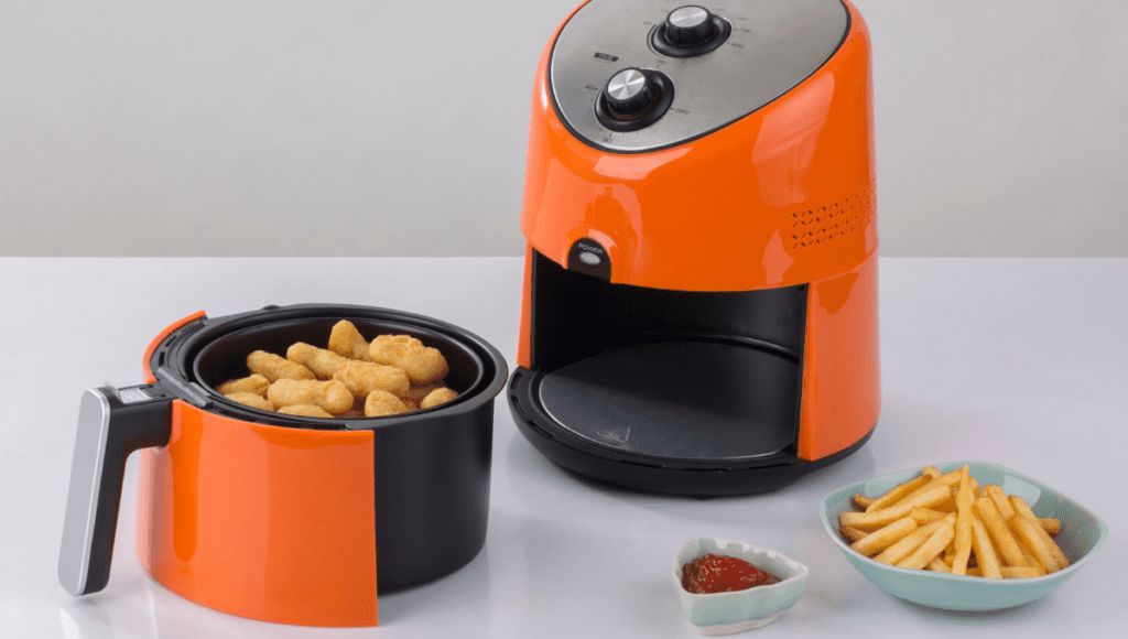 Bright orange air fryer with fries and chicken nuggets in the basket, next to a serving of fries and ketchup.