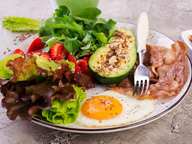 A healthy breakfast plate featuring a fried egg, crispy bacon, fresh greens, avocado, and strawberries for a nutrient-packed start to the day.