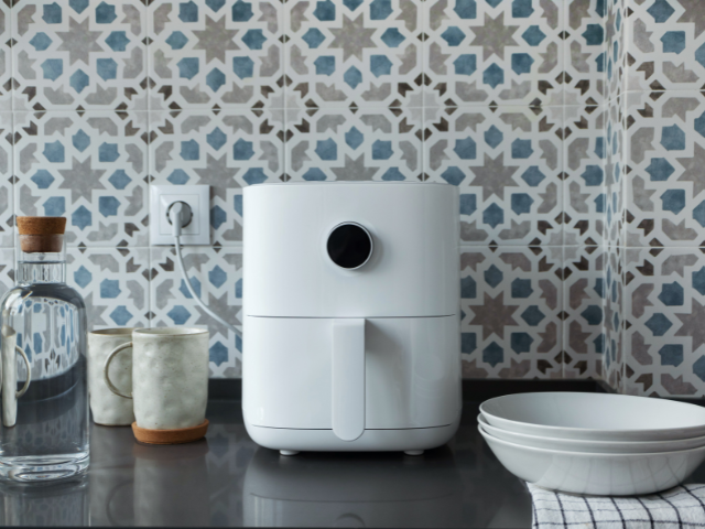 A white air fryer next to ceramic cups and bottles on a dark countertop with patterned tiles in the background.