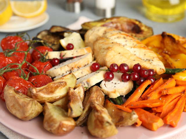 A colorful plate of roasted vegetables and sliced chicken garnished with red currants, surrounded by crispy roasted potatoes and sweet potato fries.