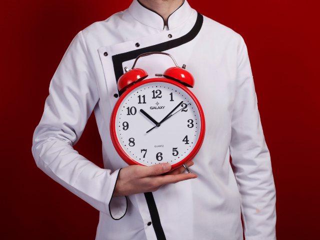 A chef holding a large red alarm clock in front of their white uniform.