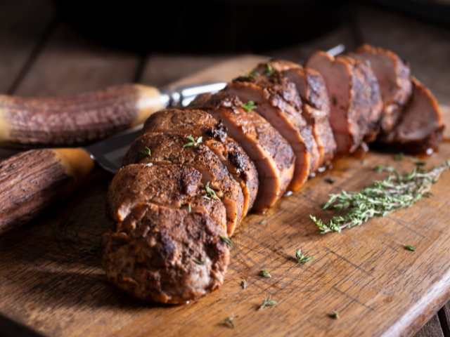 Sliced seasoned meat, cooked to perfection, on a wooden board, garnished with herbs.