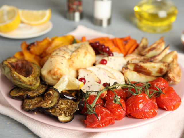 Another angle of the roasted chicken and vegetable dish, featuring cherry tomatoes on the vine and roasted eggplant, arranged artistically on a pink plate.