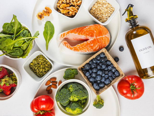 A variety of healthy foods displayed on a plate, including salmon, spinach, nuts, blueberries, broccoli, and olive oil.