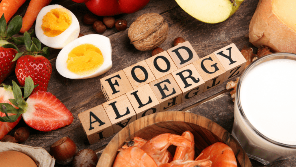 A variety of common food allergens, including strawberries, eggs, shrimp, and milk, arranged around wooden blocks spelling "FOOD ALLERGY."