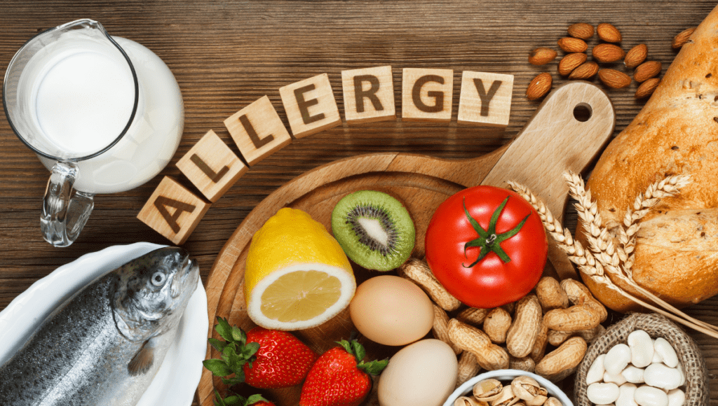 Various foods like fish, bread, eggs, nuts, and milk arranged around the word "Allergy" spelled out in wooden blocks.