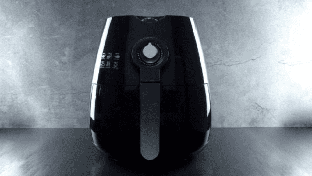 A black air fryer on a dark surface with a minimalistic gray textured wall in the background.