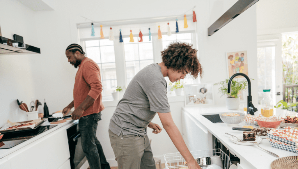 A teenager unloading the dishwasher in a bright kitchen while another person cooks in the background.