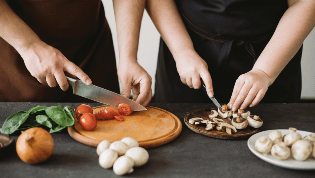 Two people cutting vegetables, one slicing tomatoes and the other chopping mushrooms.