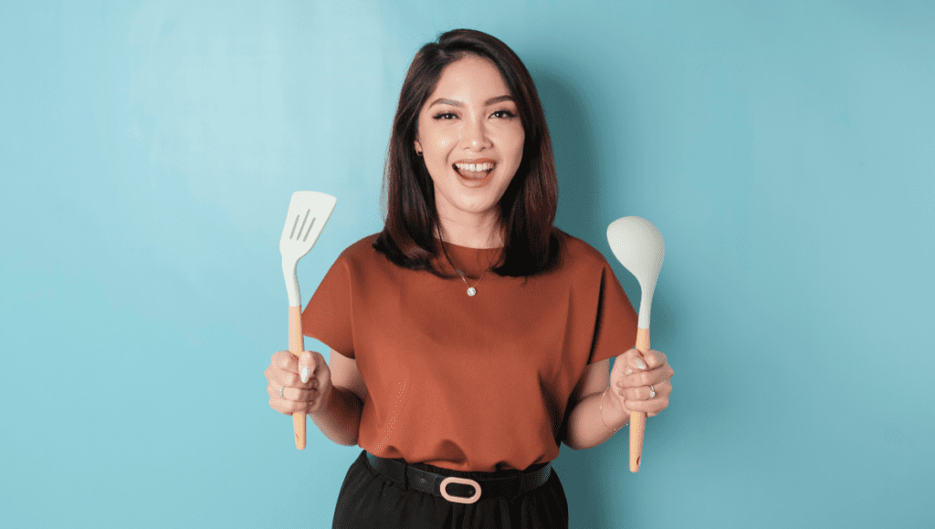 A cheerful woman holding a spatula and spoon, standing against a light blue background.