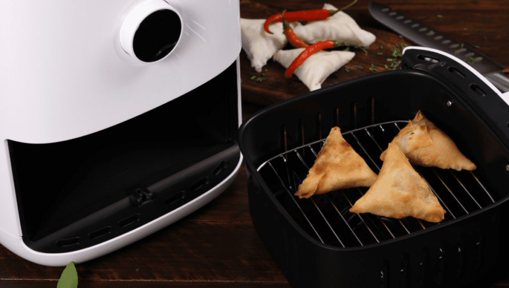 Three golden-brown samosas sitting inside an air fryer basket, with uncooked samosas and red chili peppers in the background.