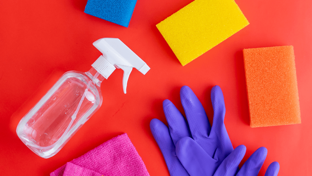 A cleaning setup with various sponges, a spray bottle, purple gloves, and a cloth on a bright red background.