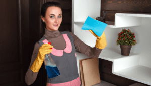 A woman wearing cleaning gloves holding a spray bottle and sponge, standing in a home setting ready to clean.