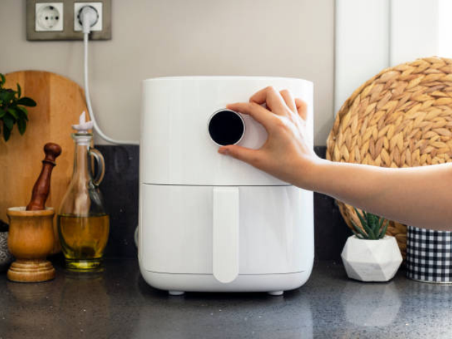 A person turning the dial on a sleek, white air fryer on a kitchen counter, surrounded by cooking tools like a bottle of olive oil.