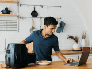 A man preparing food using an air fryer while looking at a laptop.