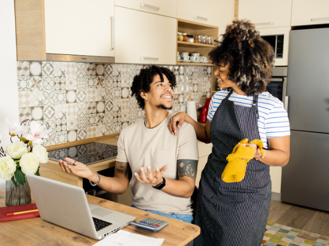 A man sits at a kitchen counter with a laptop while a woman stands beside him wearing an apron and holding an oven mitt.