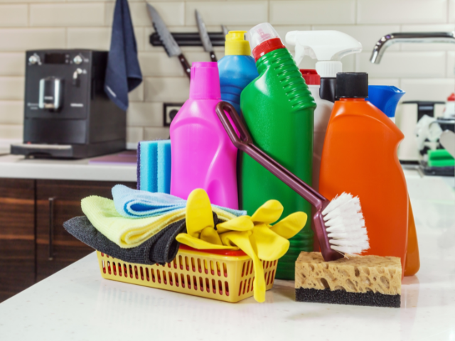 A basket filled with various colorful cleaning supplies including bottles, gloves, and sponges on a kitchen counter.