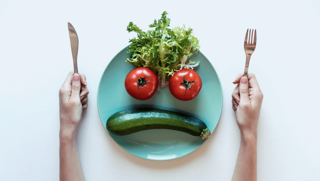 A plate arranged with two tomatoes, lettuce, and a zucchini to form a sad face, with hands holding a knife and fork on either side.