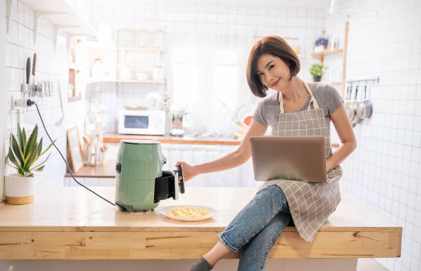 Woman sitting on a kitchen counter using a laptop while preparing food with an air fryer.